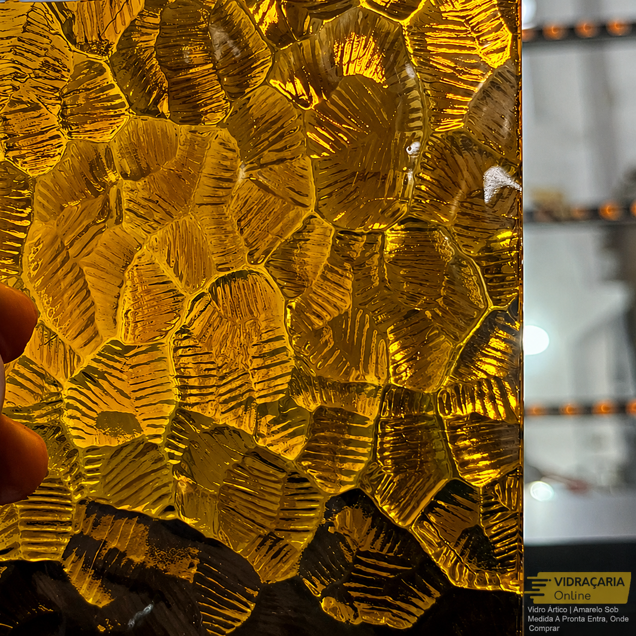 Placa de vidro artesanal com peças amareladas em formato de folhas geométricas, criadas por encaixe, com iluminação interna que realça o relevo. Ao fundo, uma loja com vitrines desfocadas.